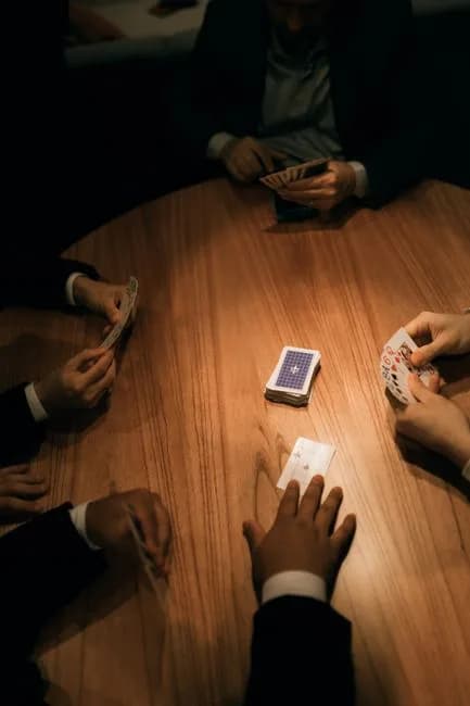 A group of adults playing a card game around a wooden table in moody lighting, representing a teen patti session
